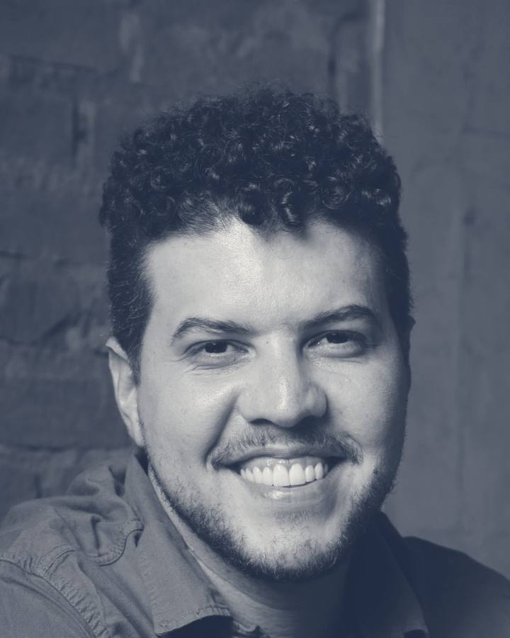 Marcus Vinícius, revisor de textos, sorrindo, com as mãos cruzadas segurando uma caneta ao lado do notebook